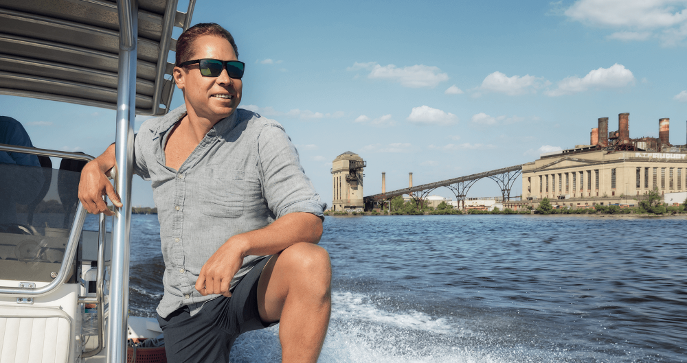 Wearing sunglasses, Zack Hoisington '07 speeds along the Delaware River in a boat near Philadelphia and Camden. White water is splashing up from the sides and there is a bridge and an older beige building in the background.