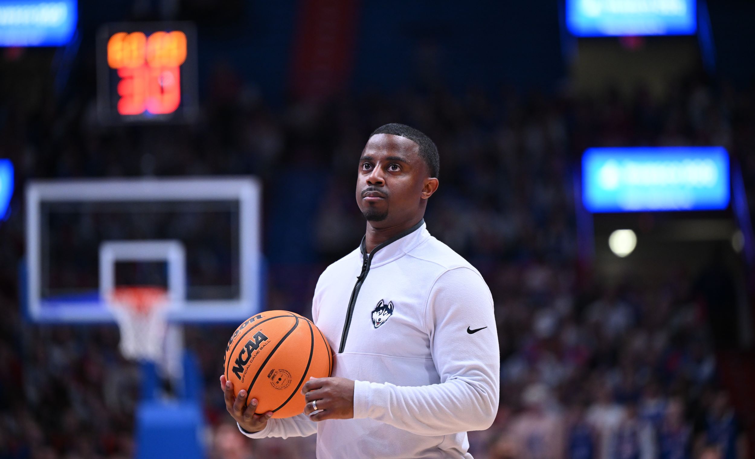 Mathew Johnson '13, an assistant coach for UConn men's basketball, holds a ball and looks up from the court while wearing a white UConn Huskies quarter-zip pullover.