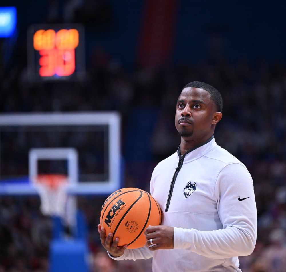 Mathew Johnson '13, an assistant coach for UConn men's basketball, holds a ball and looks up from the court while wearing a white UConn Huskies quarter-zip pullover.