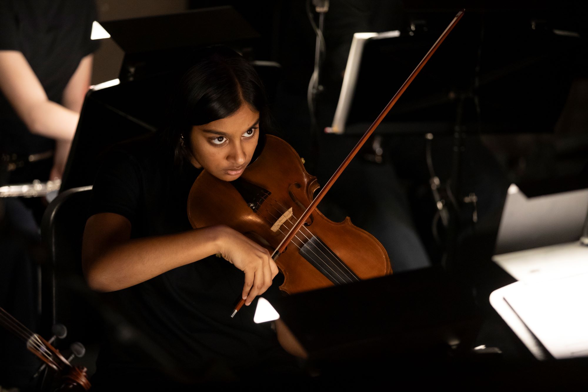 In the orchestra pit, Arya Vishwakarma '25 looks up intently at the conductor, who is not in the frame, as she moves her bow across thew strings of her viola.