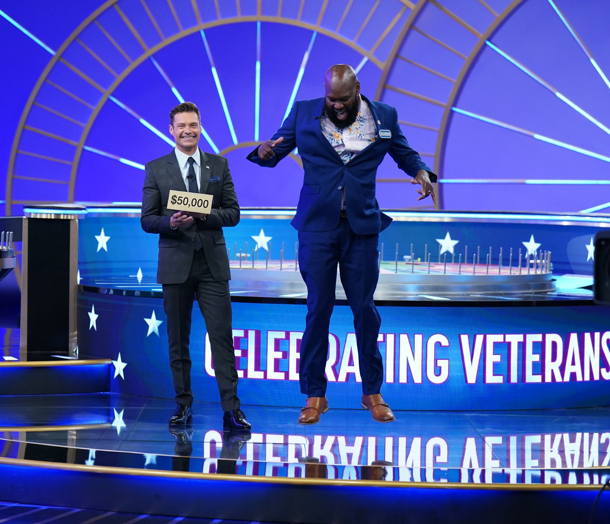 Brandon Shields '03 jumps in the air on set of Wheel of Fortune after being presented with his $50,000 prize from host Ryan Seacrest, standing to Brandon's right.