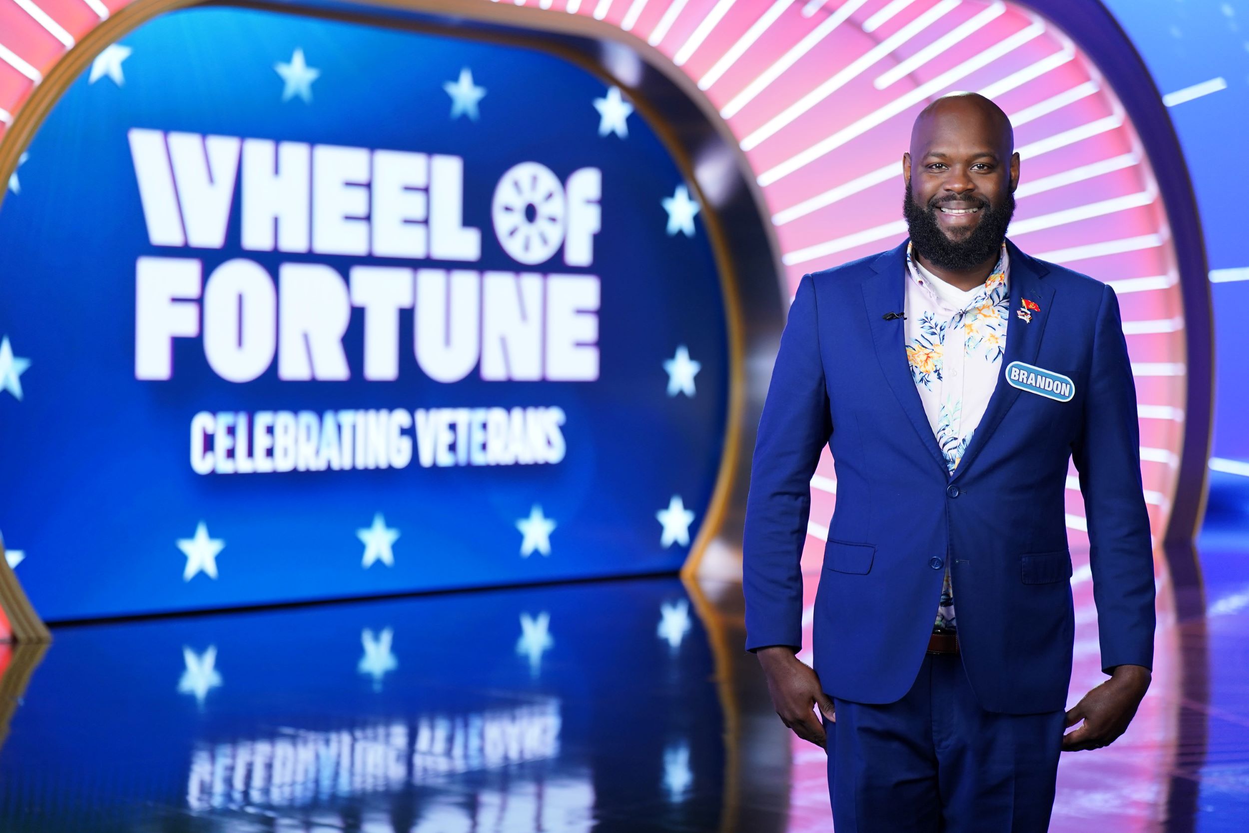 Brandon Shields '03, wearing a blue suit and a white shirt, stands on the set of Wheel of Fortune in a publicity shot from his appearance on the show during Veterans Week.