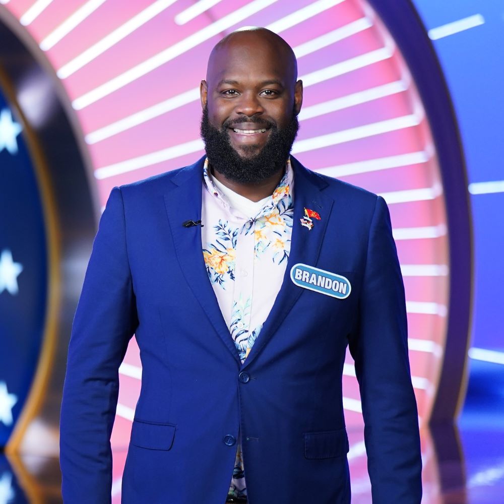 Brandon Shields '03, wearing a blue suit and a white shirt, stands on the set of Wheel of Fortune in a publicity shot from his appearance on the show during Veterans Week.