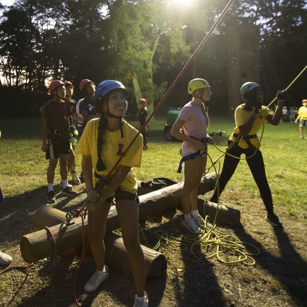 New Second Formers participating at the Ropes Course, standing on the ground while they hold ropes attached to classmates who are climbing the course.