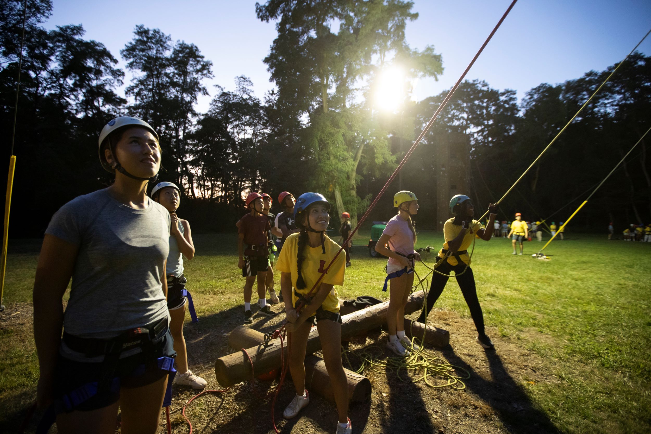 New Second Formers participating at the Ropes Course, standing on the ground while they hold ropes attached to classmates who are climbing the course.