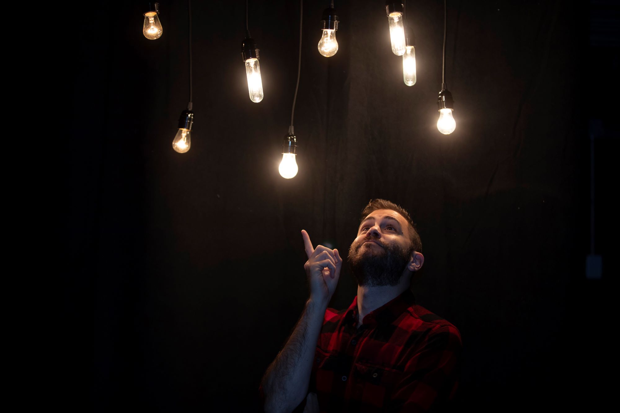 Wearing a red and black checked flannel shirt, in a dimly lit room, Jared Goldenberg points up to the hanging lit bulbs above his head.