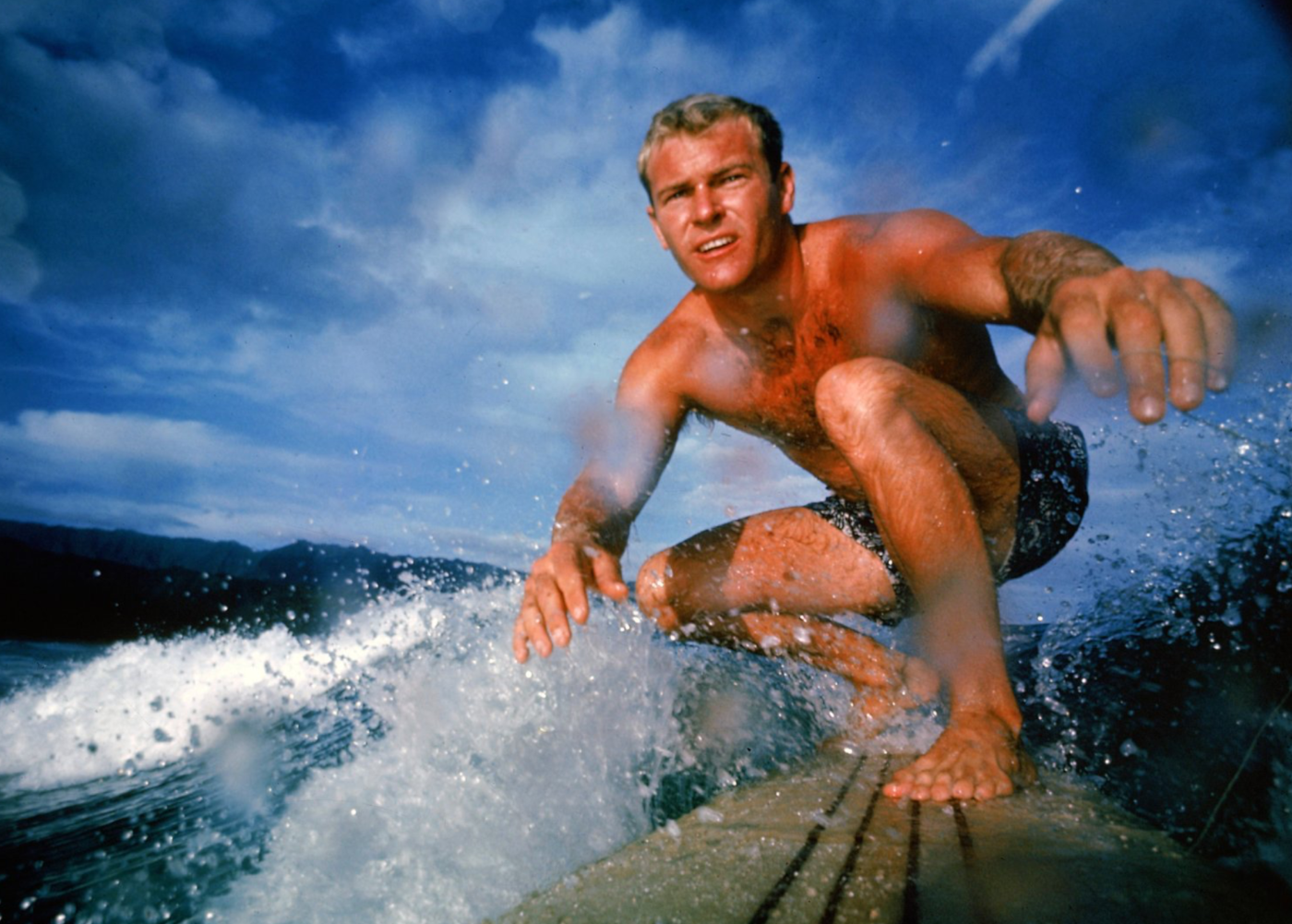 In a color photo from 1963m Nick Beck '58 rides a wave on his light board in a crouched position.