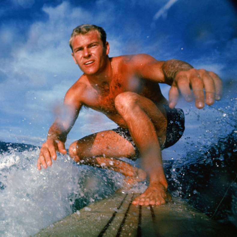 In a color photo from 1963m Nick Beck '58 rides a wave on his light board in a crouched position.