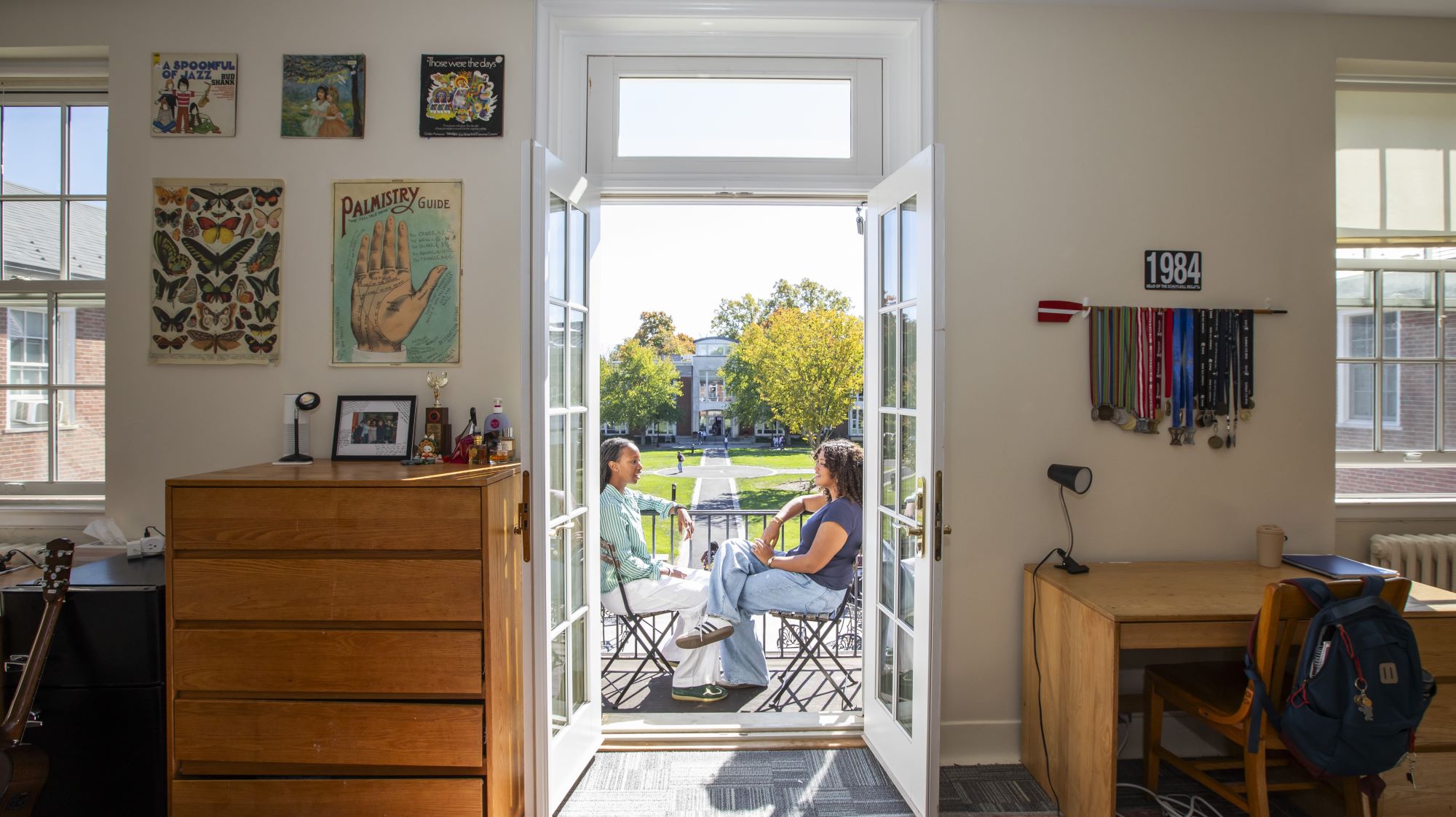 From a view inside their room in Macpherson House, two girls chat on their room's veranda, visible through the open French doors, overlooking Noyes Quad.
