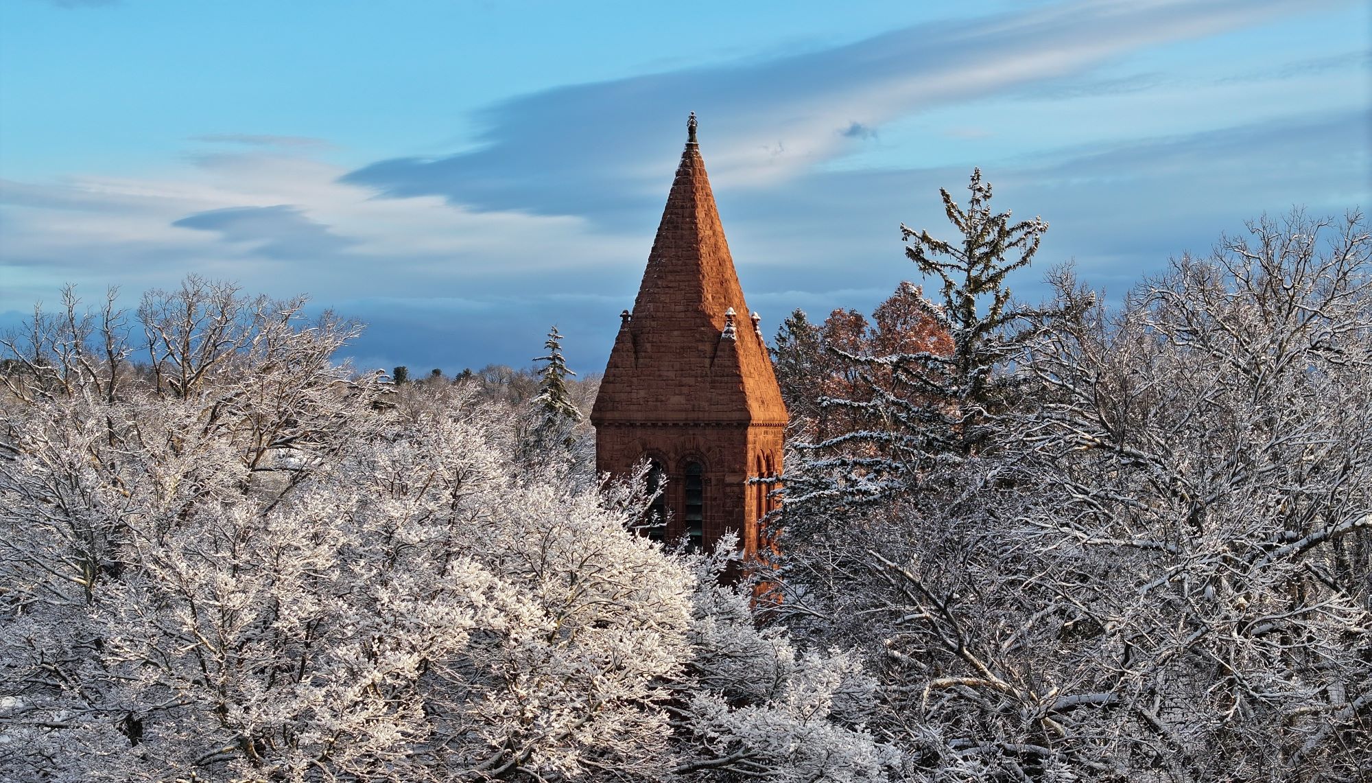 The brownstone steeple of Edith Memorial Chapel rises above the tops of snow-laced trees in an aerial shot.