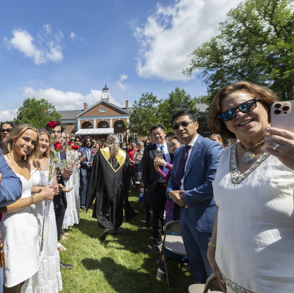 This photo is shot from the perspective of someone marching in the Commencement processional, with attendees on both sides of the pathway looking at the photographer with enthusiasm.