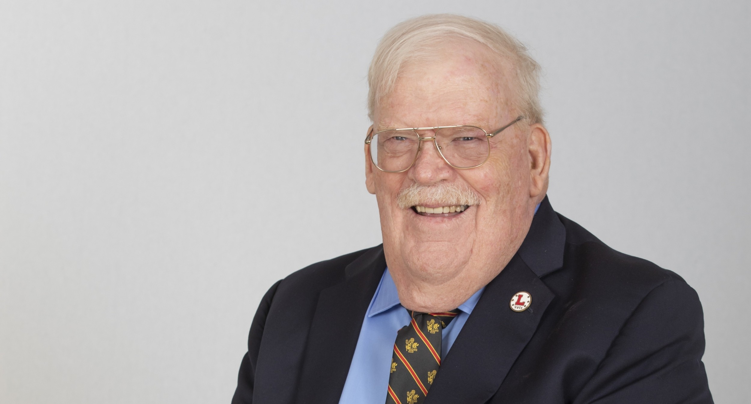 Portrait of Matt Dominy '65 wearing a blue blazer with a Lawrenceville seal and a black, red, and gold Lawrenceville tie over a light blue shirt.