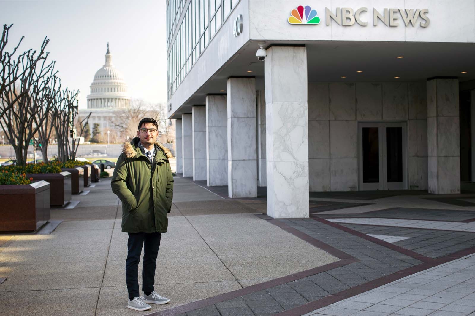 Wearing a winter parka, Ghael Fobes is standing outside the Washington Bureau of NBC News, with the U.S. Capitol building in the distant background.
