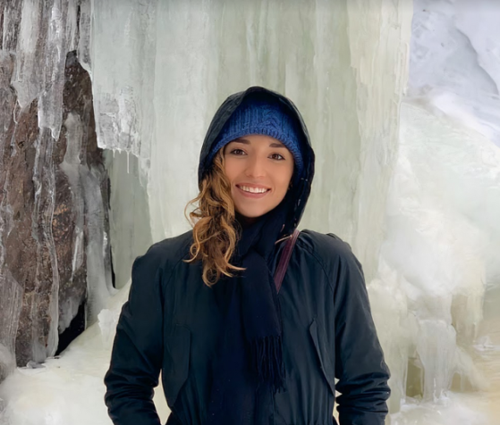 Hannah-Marie Garcia-Ladd '15, wearing a woolen hat and a heavy winter coat with her hood up, stands smiling before a cascade of ice in Alaska