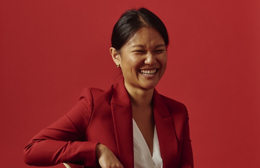Karen Hao is sitting in a chair and laughing. She is wearing an orange suit with a white blouse in front of an orange background.