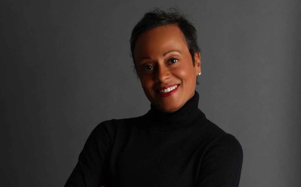 Monica Williams '91, wearing a black turtleneck, had her arms folded and she is smiling in front of a dark background.