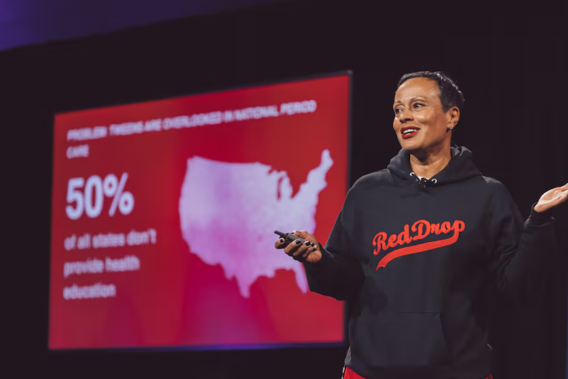 Monica Williams '91, wearing a black hoodie with "Red Drop" emblazoned in red script, speaks to an audience with a sales graphic on a screen behind her.