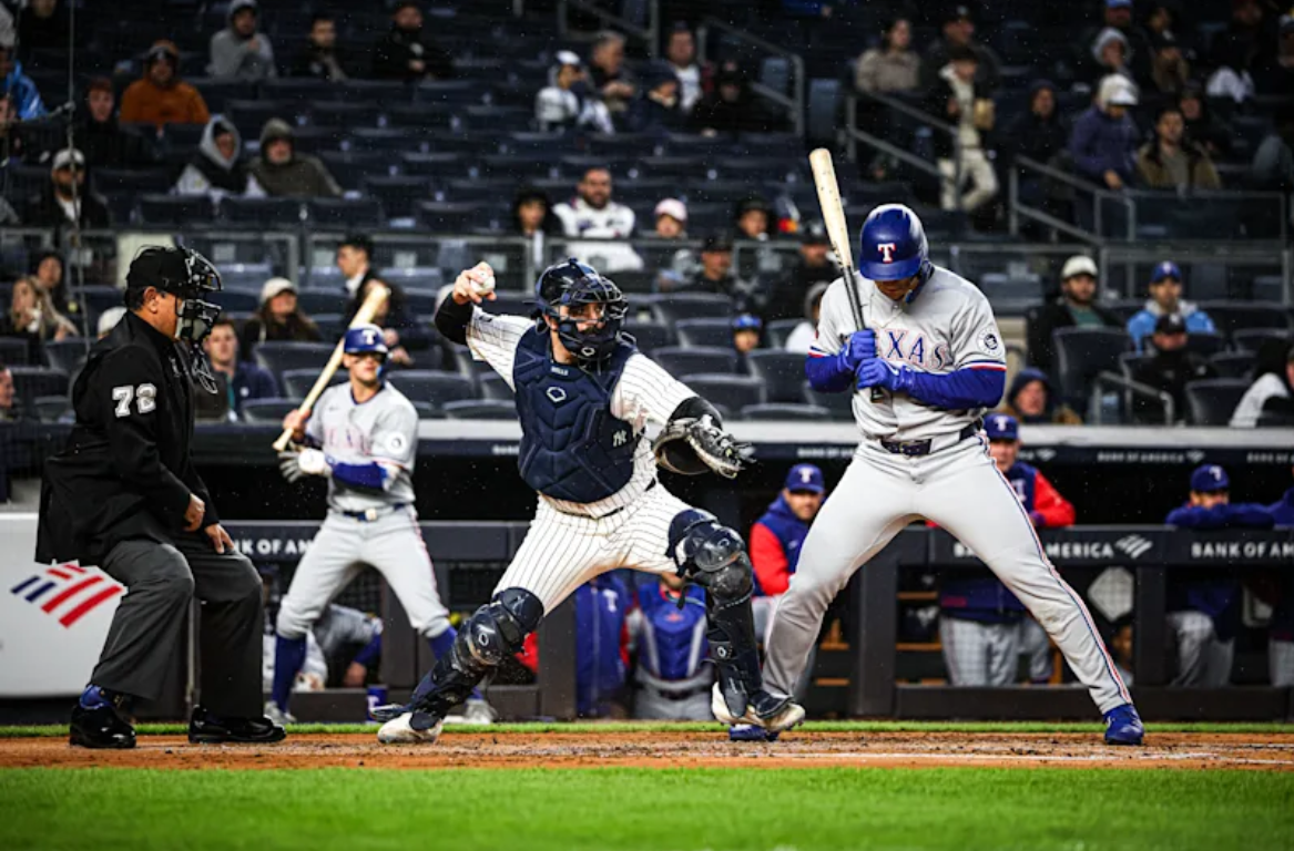 A New York Yankees catcher has his arm cocked to throw the baseball as a Texas Rangers batter remains in his batting stance.