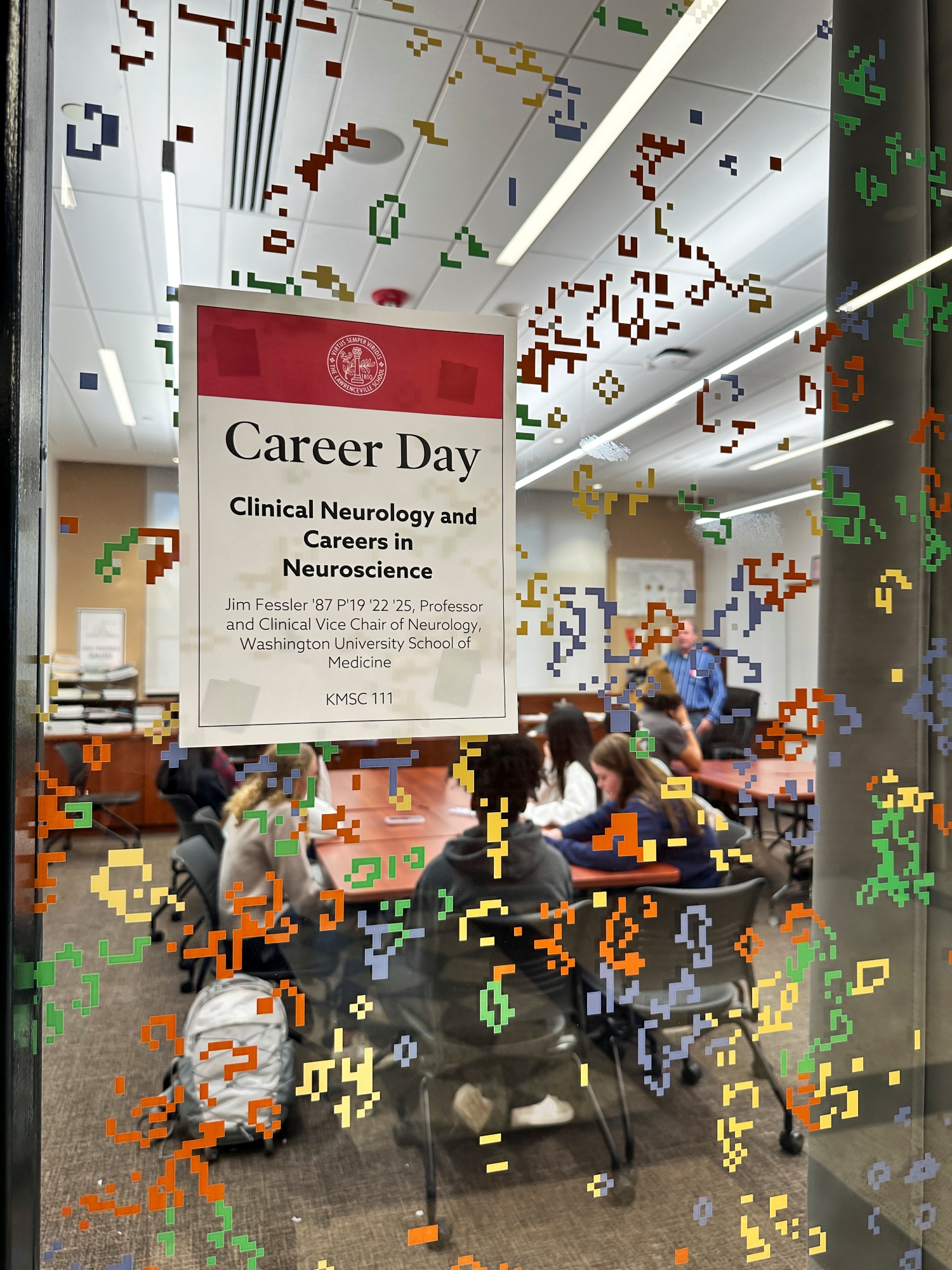 A glass door covered with colorful stickers and a Career Day sign partially obscure the classroom inside, filled with students and an alumnus talking about his career.