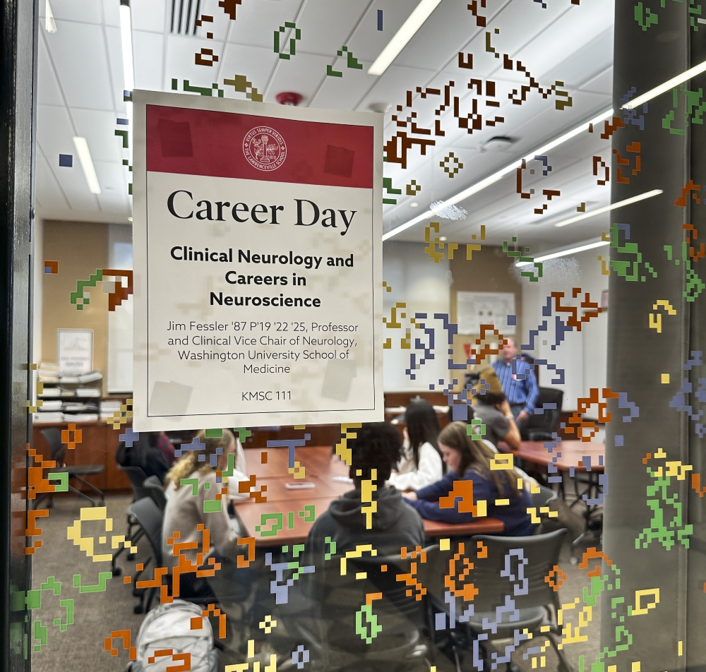 A glass door covered with colorful stickers and a Career Day sign partially obscure the classroom inside, filled with students and an alumnus talking about his career.