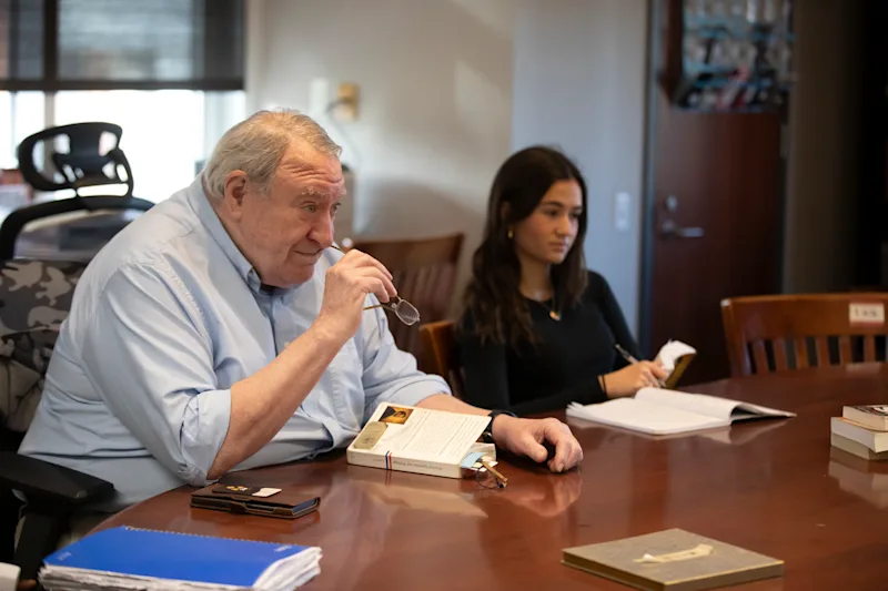Teacher Regan Kerney, sitting at a brown Harkness table, holds his eyeglasses at face level while looking at a student we cannot see. A girl student sits to Kerney's left at the table.