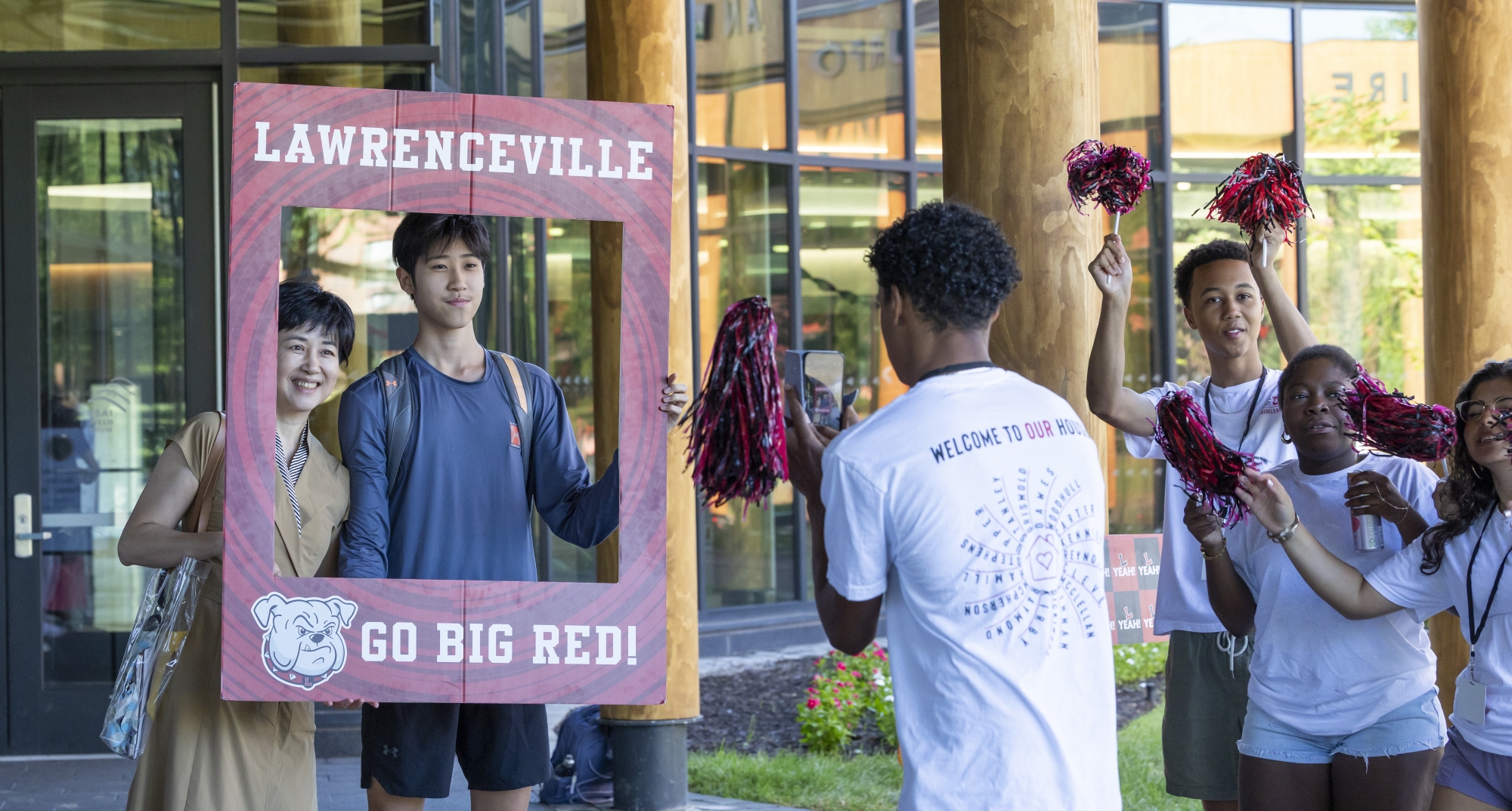 A new student and his mother pose inside a red "Lawrenceville - Go Big Red!" frame while another student takes their photo.