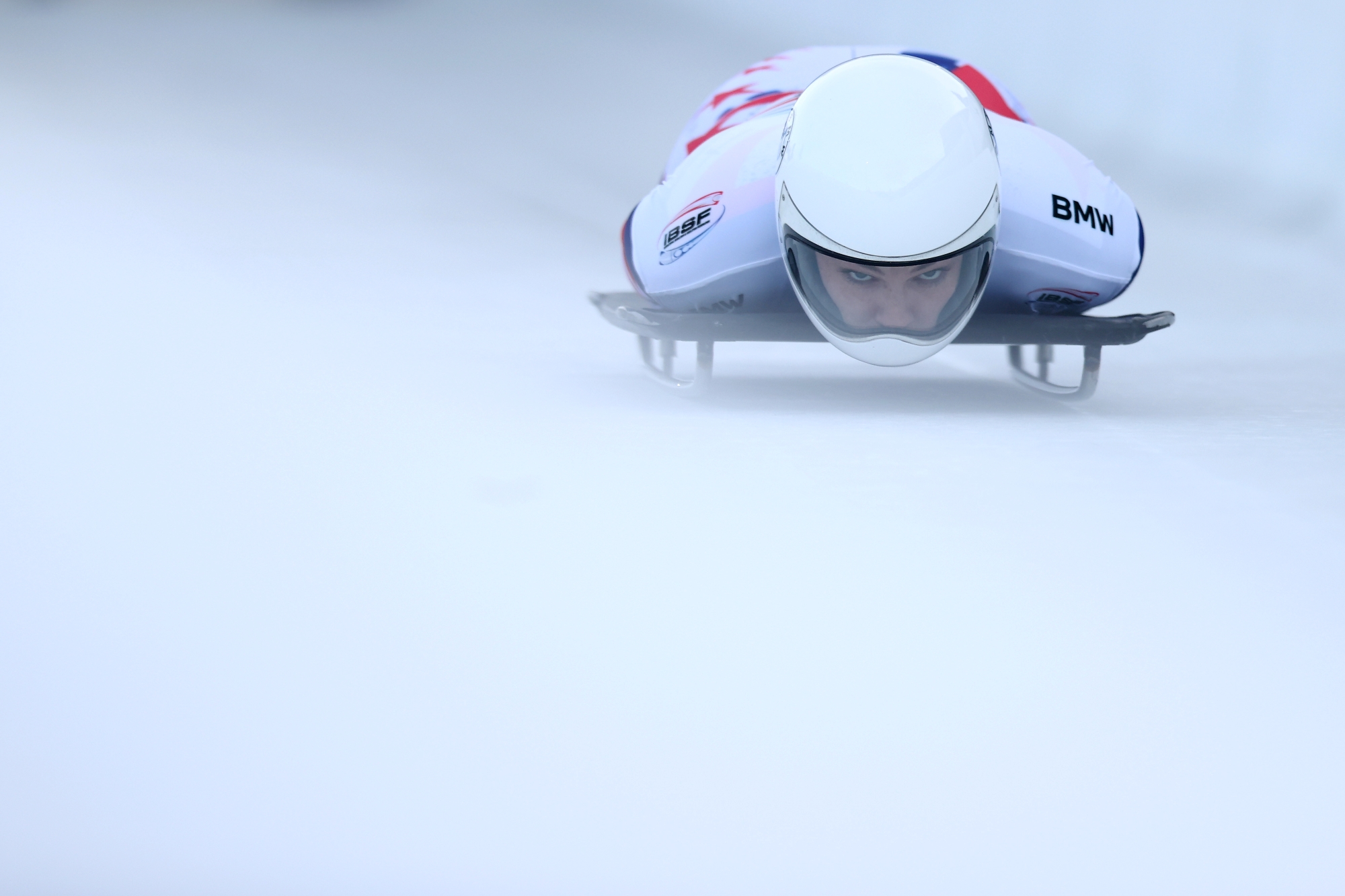 Lying flat on her stomache with arms tucked, Kelly Curtis stares straight ahead through the visor of her white helmet as she speeds downhill on her skeleton aparatus. Her speed is perceptible through the still image.