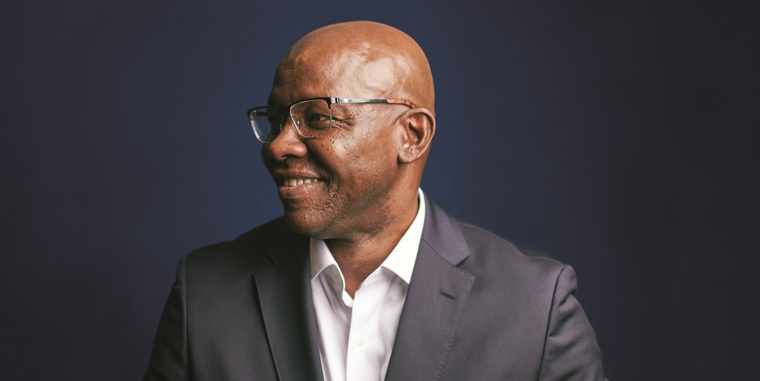 Garry D. Howard '77 is wearing a dark blue suit and a white shirt open at the neck, and a white pocket square. He is wearing glasses and smiling, while looking outside the camera frame to the viewer's left.