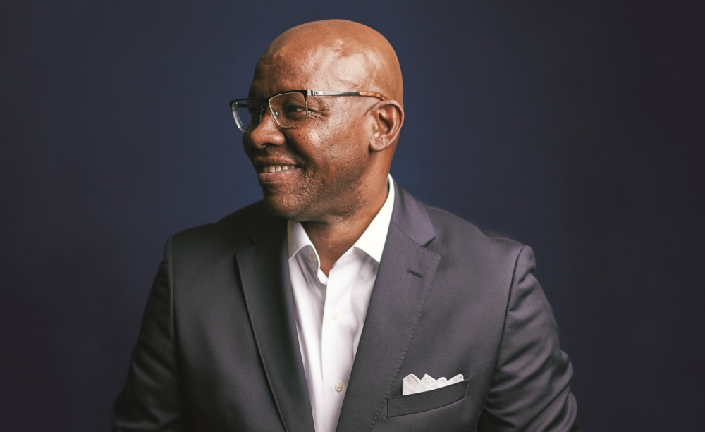 Garry D. Howard '77 is wearing a dark blue suit and a white shirt open at the neck, and a white pocket square. He is wearing glasses and smiling, while looking outside the camera frame to the viewer's left.