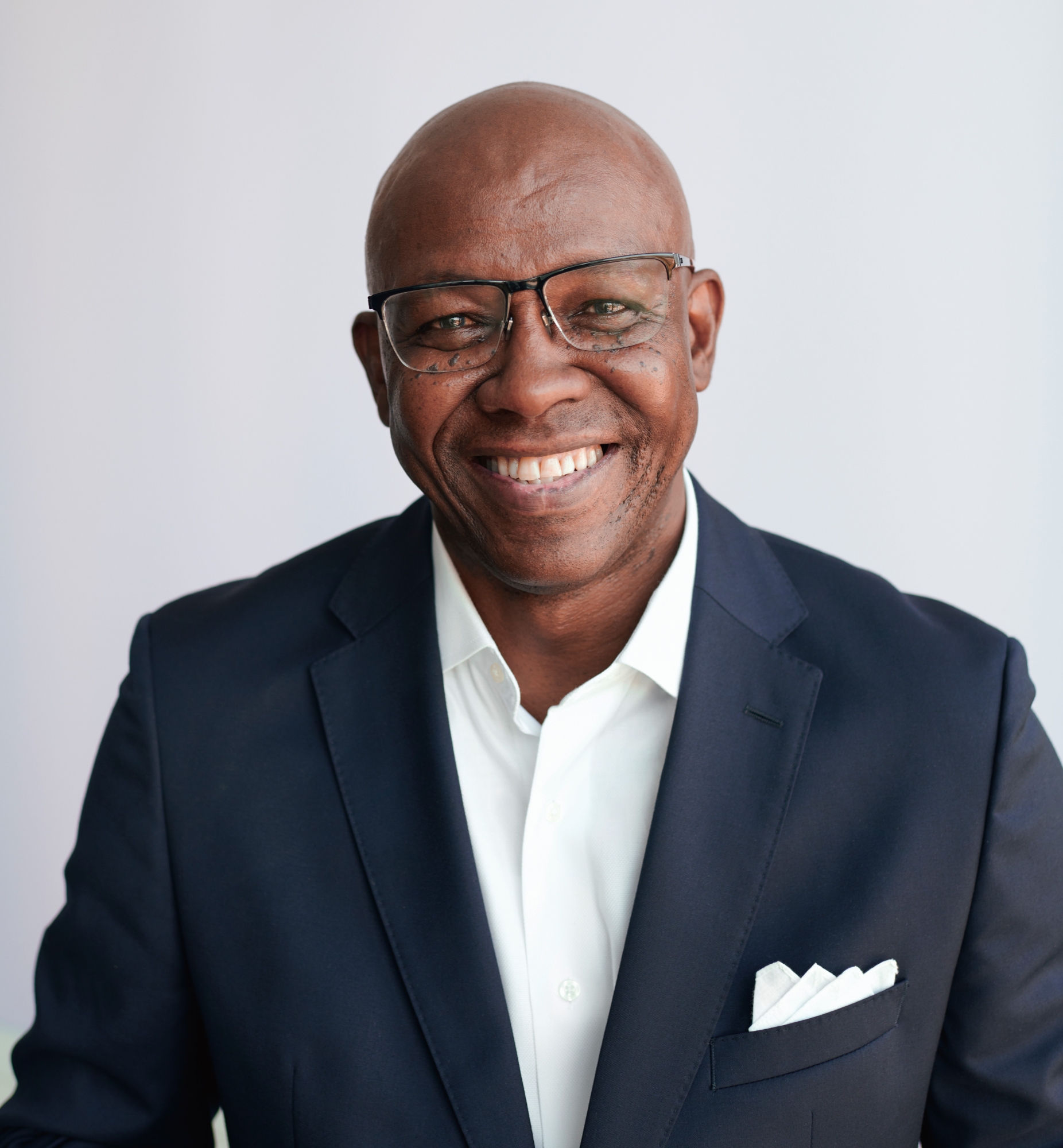 Garry D. Howard '77 is wearing a dark blue suit and a white shirt open at the neck, and a white pocket square. He is wearing glasses and smiling, while looking toward the camera.