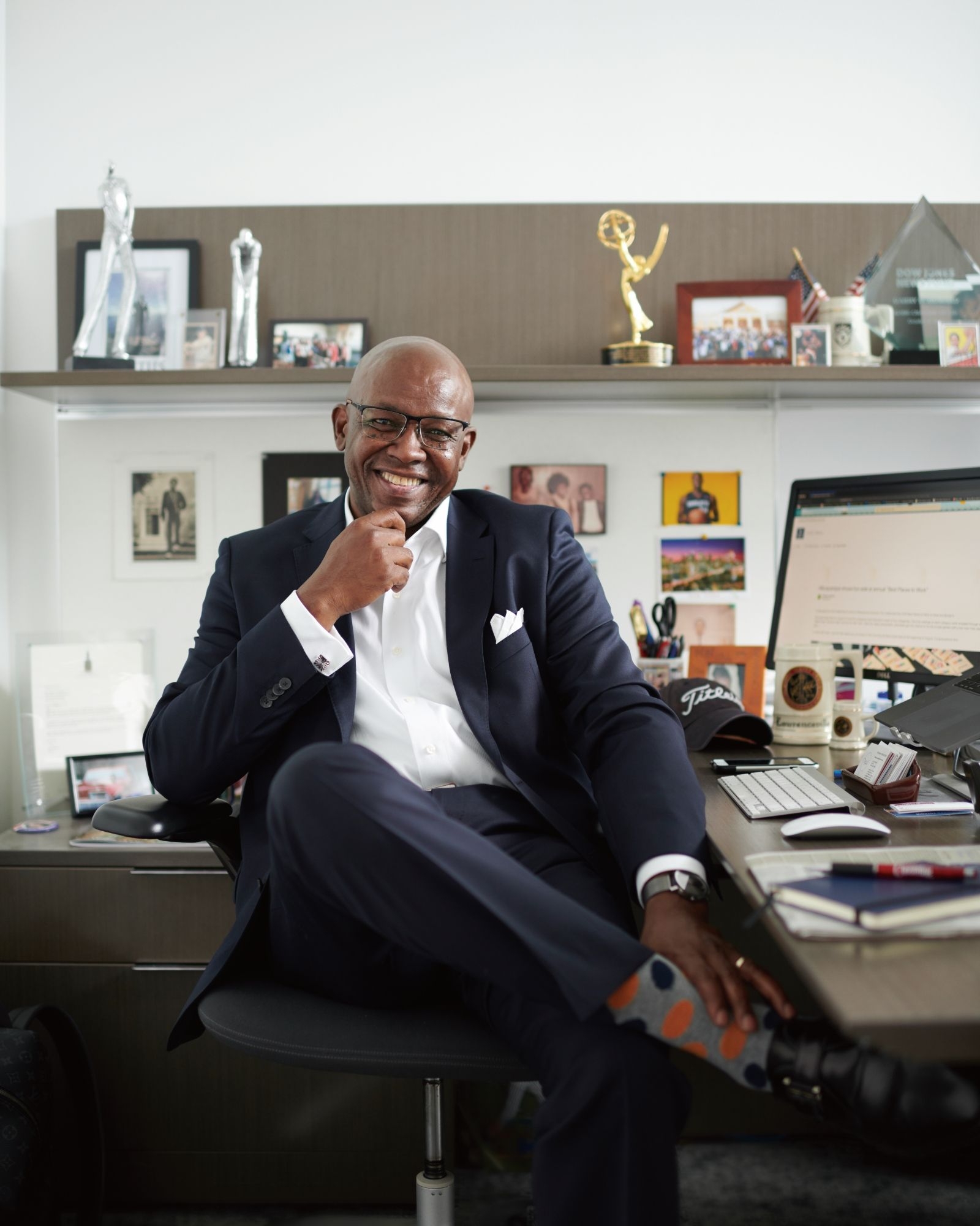 Garry D. Howard '77 is wearing a dark blue suit and a white shirt open at the neck, and a white pocket square. Sitting at the desk in his office, he is wearing glasses and smiling, while looking toward the camera.
