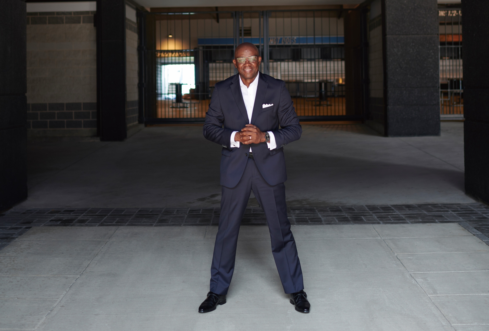 Garry D. Howard '77 is wearing a dark blue suit and a white shirt open at the neck, and a white pocket square. Standing with his hands clasped inside the NFL stadium of the Carolina Panthers, he is wearing glasses and smiling, while looking toward the camera.