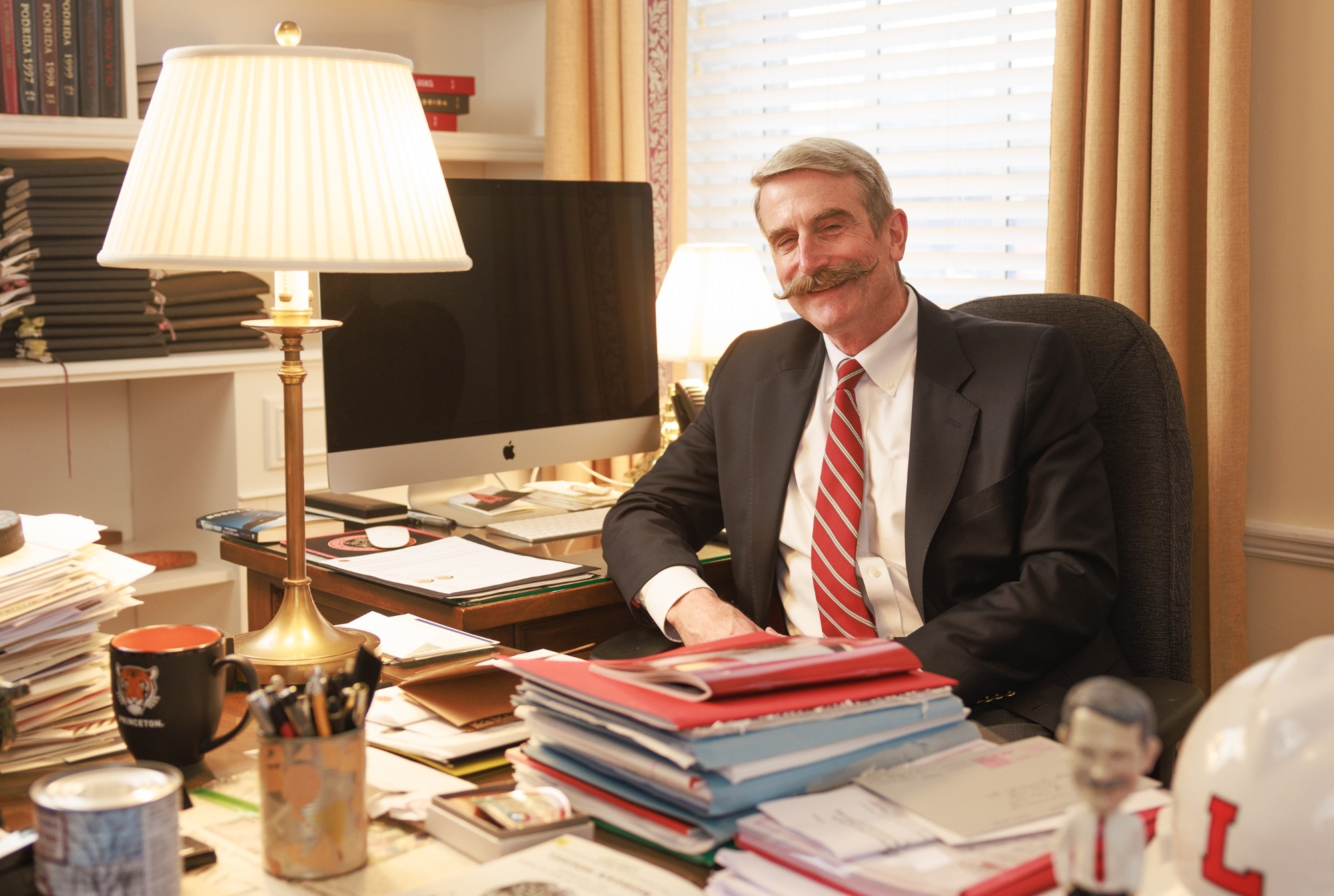 Steve Murray sitting at his office desk, smiling. He is wearing a blue blazer, white oxford button-down short, and a red and black tie. His desk is covered with papers and notebooks and many mementos he has collected over the years.