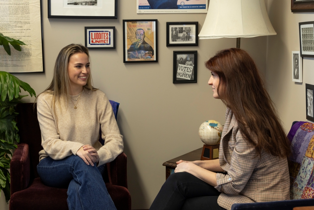 Leila Campbell '26, a student, is wearing a beige sweater and jeans and is smiling as she talks to history teacher Hazel Baldwin-Kress, who we see in profile as she looks at Leila.