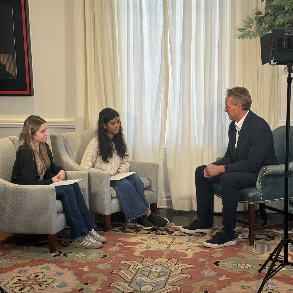 Sen. Jeff Flake, wearing a dark suit, sits in a chair with his hands clasped by his knees, while he talks to two girl seated students in an interview.