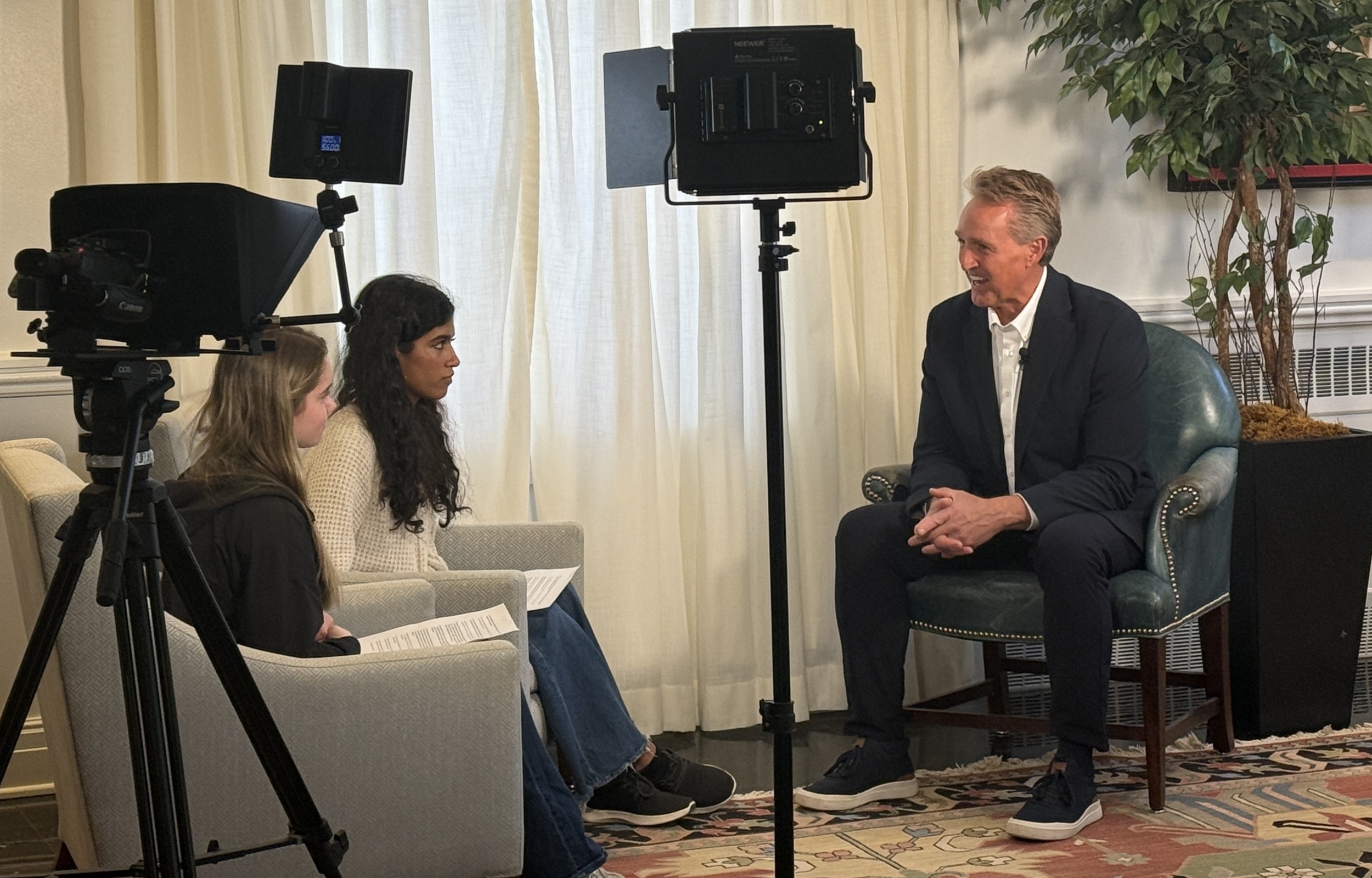 Sen. Jeff Flake, wearing a dark suit, sits in a chair with his hands clasped by his knees, while he talks to two girl seated students in an interview.