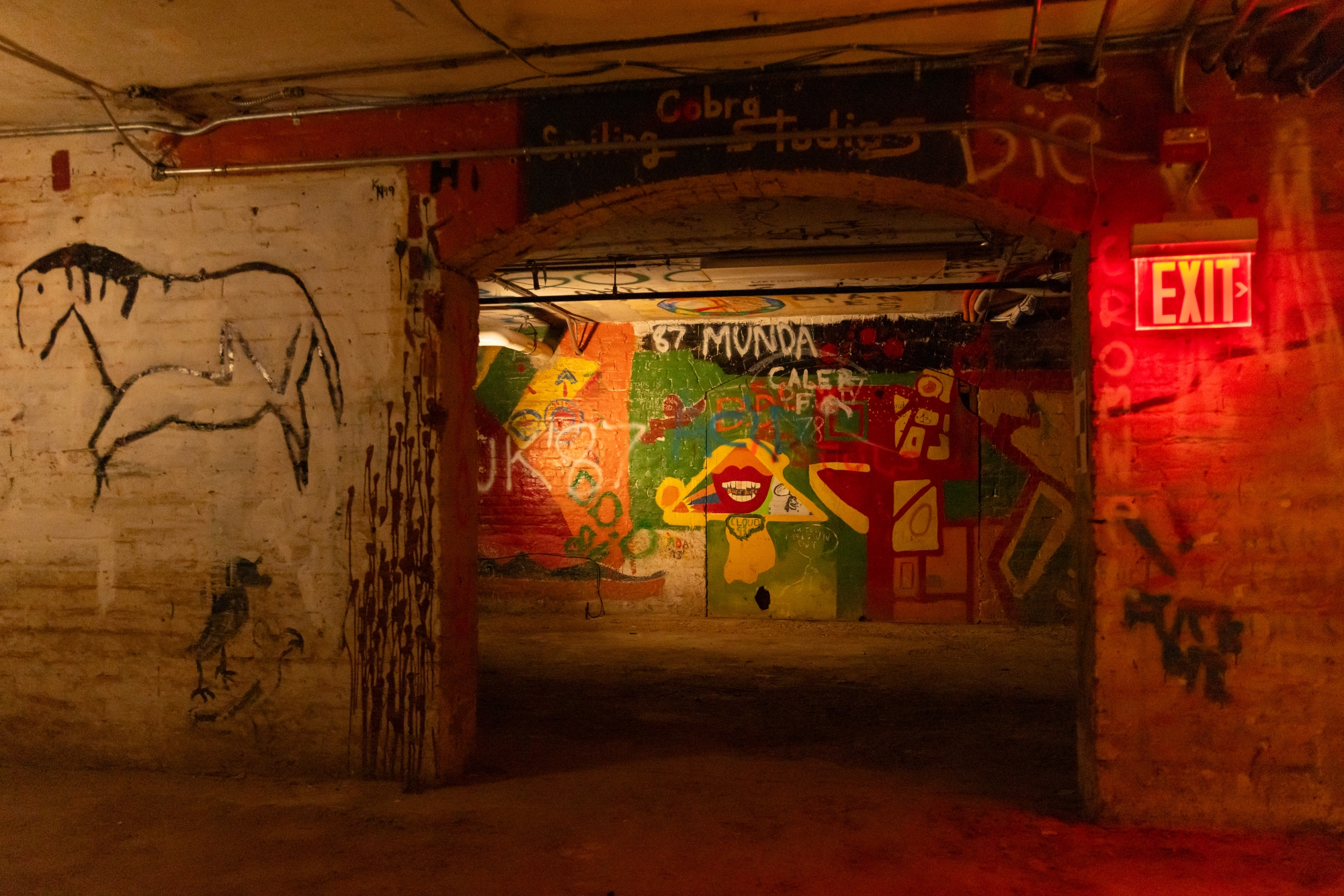 Looking through a brick archway in the basement of Memorial Hall, there is a colorful, green, orange, and yellow mural painted on the wall in the next room, with red lips being the focal point.
