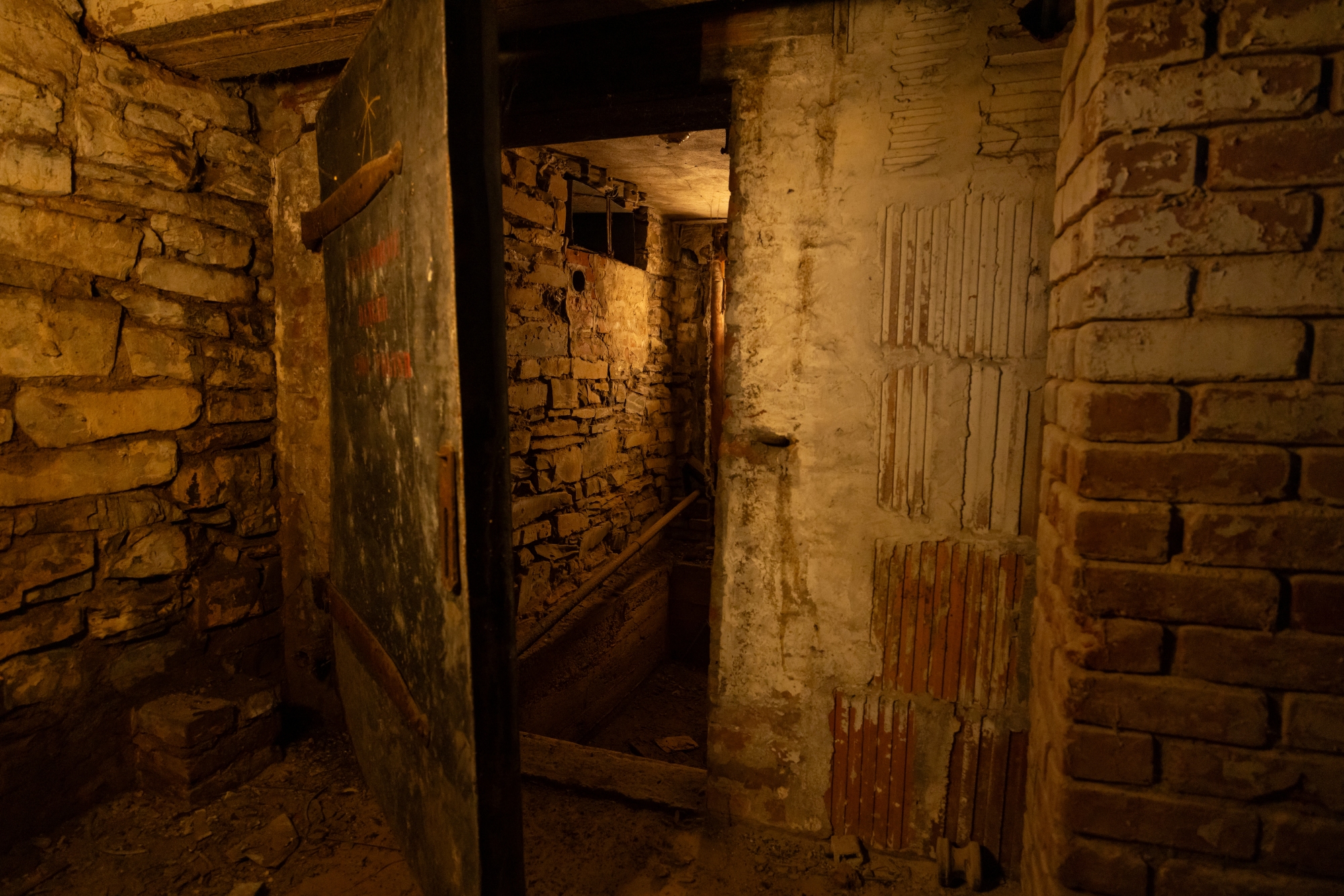 A lightly open steel door reveals a slightly lit area in the basement of Haskell House that used to serve first as an ice house and then as housing for an electrical generator. Both have long since passed into obsolescence. The room is made from stone, brick, and terra cotta-like blocks.