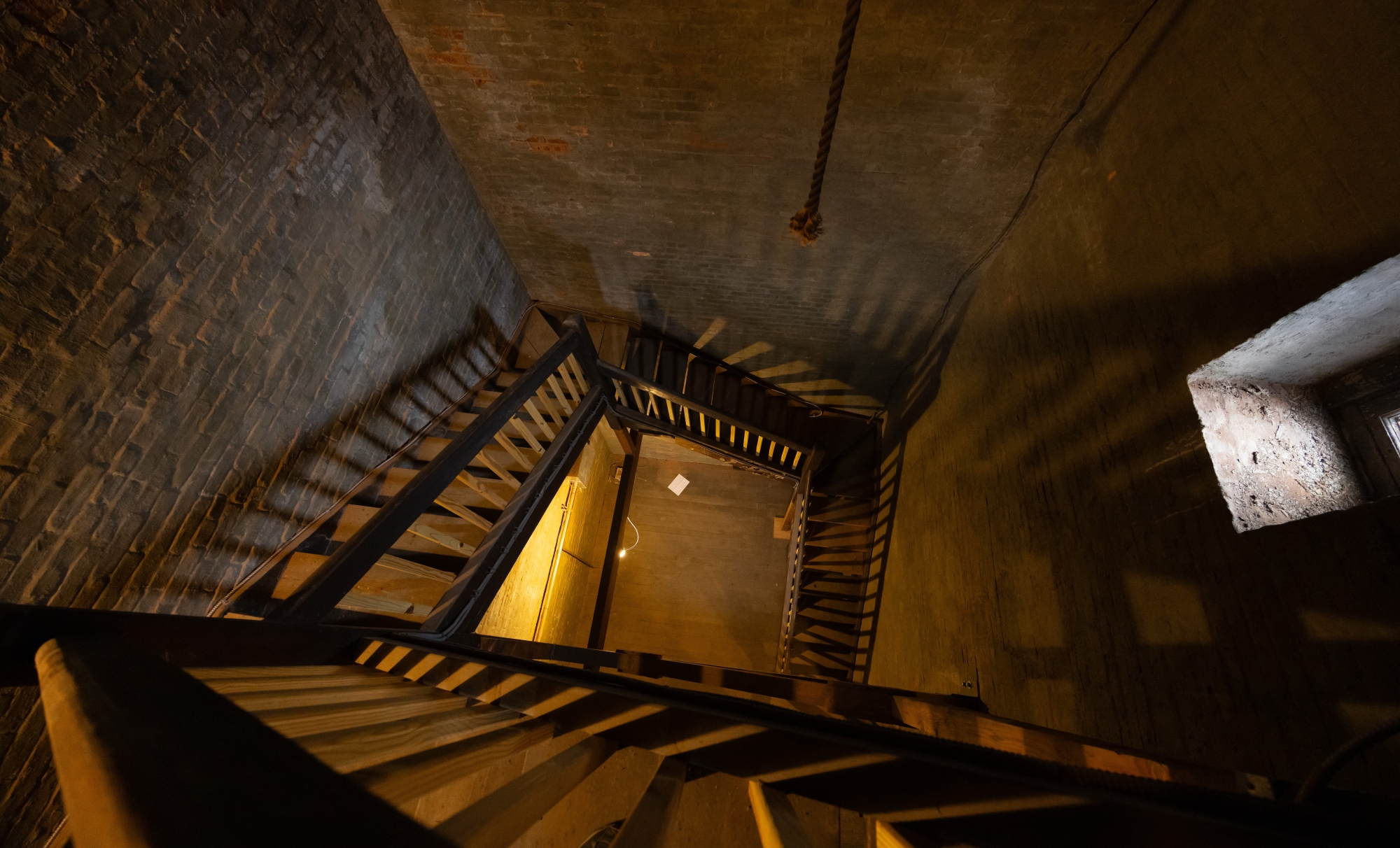 Looking down from the belfry of the Edith Memorial Chapel bell tower, there are flights of stairs against each of the tower's four walls. At the bottom, there is a wooden landing, far below.