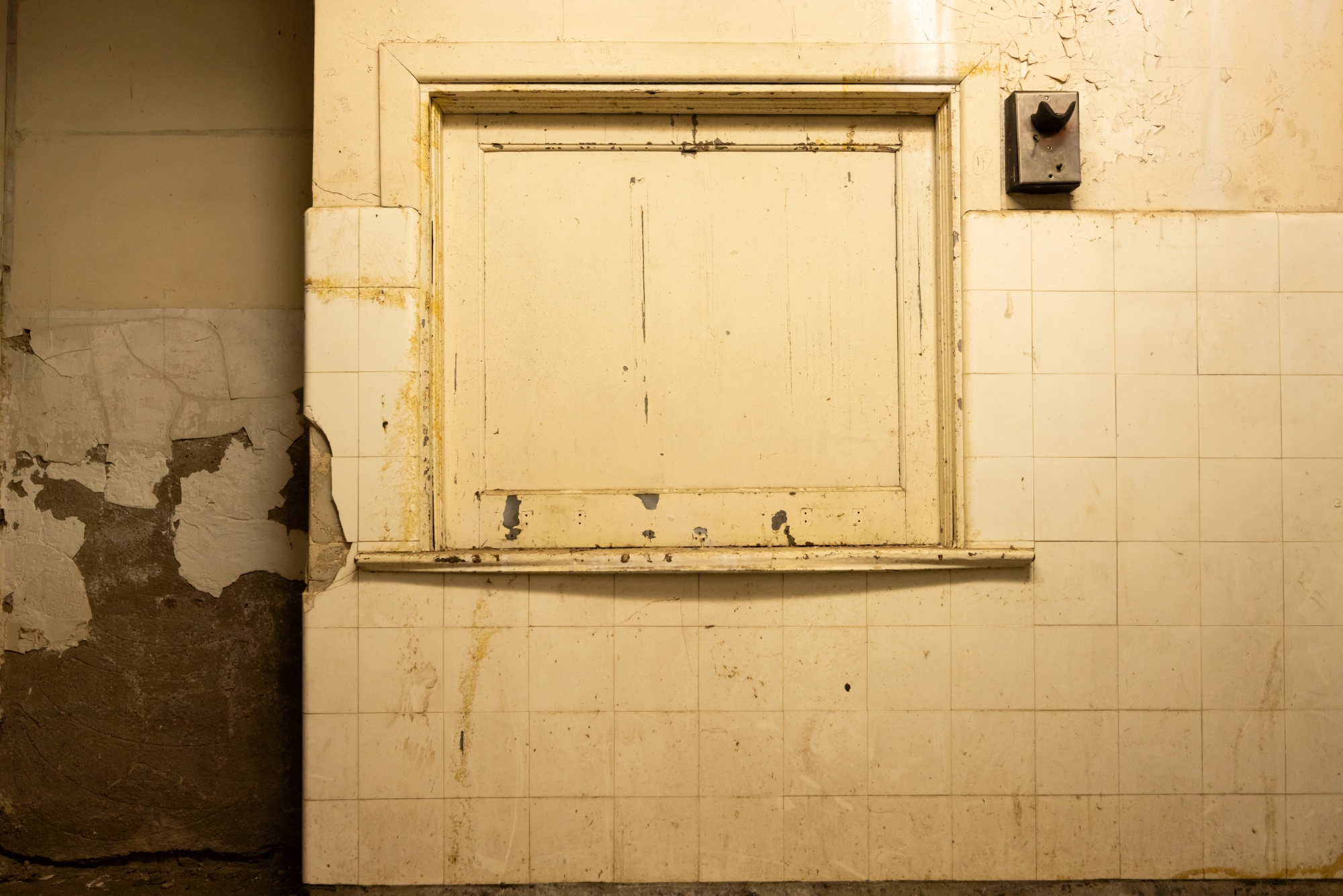 In the old infirmary, which served the School from 1929-76, a closed dispensary window is situated within a wall covered with sterile white tiles that have been dirtied over the years from inactivity.