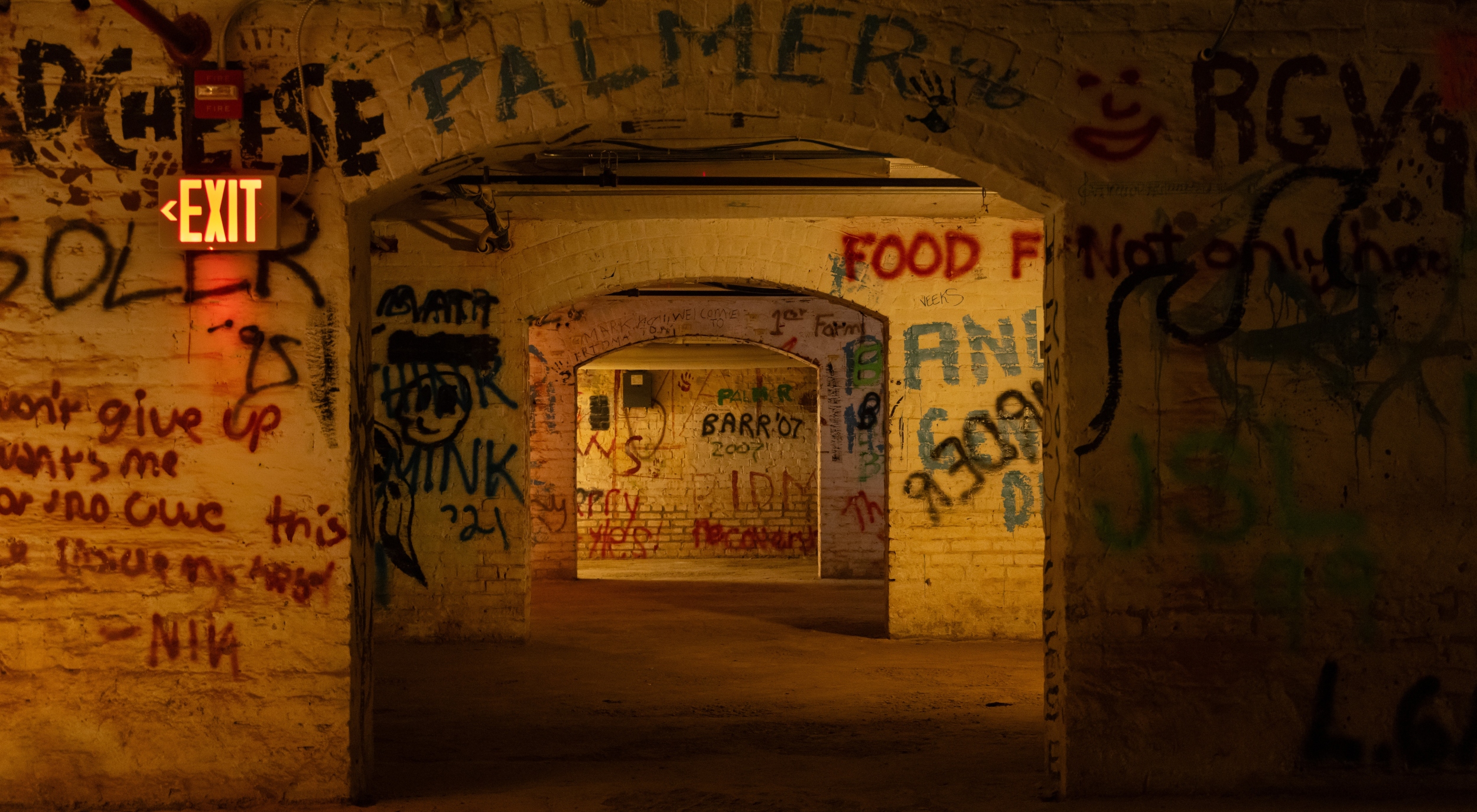Looking through a series of three brick archways in the dark basement of Memorial Hall. The walls are white but most surfaces are sprayed with colorful graffiti.