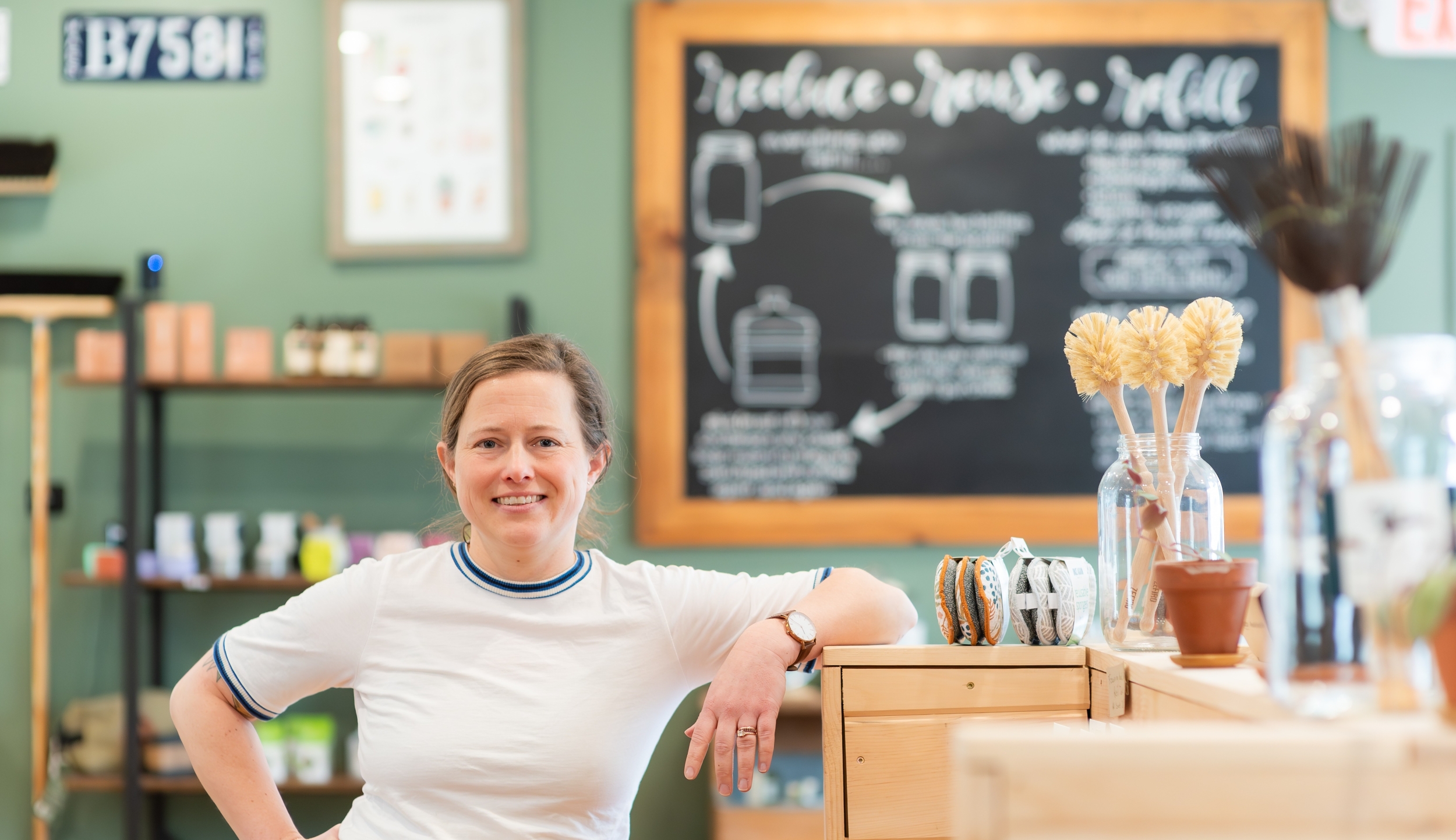 Julia Traggorth '05 is wearing a white ringer T-shirt with dark blue banding and brown corduroy pants. She is smiling as she leans against a shelf stocked in her refill store.
