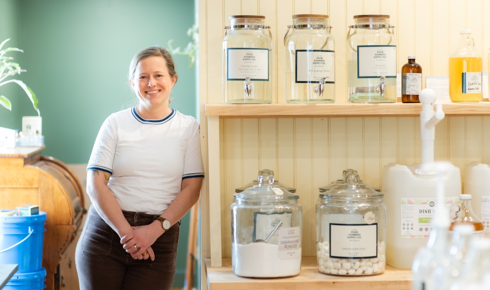 Julia Traggorth '05 is wearing a white ringer T-shirt with dark blue banding and brown corduroy pants. She is smiling as she leans against a shelf stocked with glass jars in her refill store.