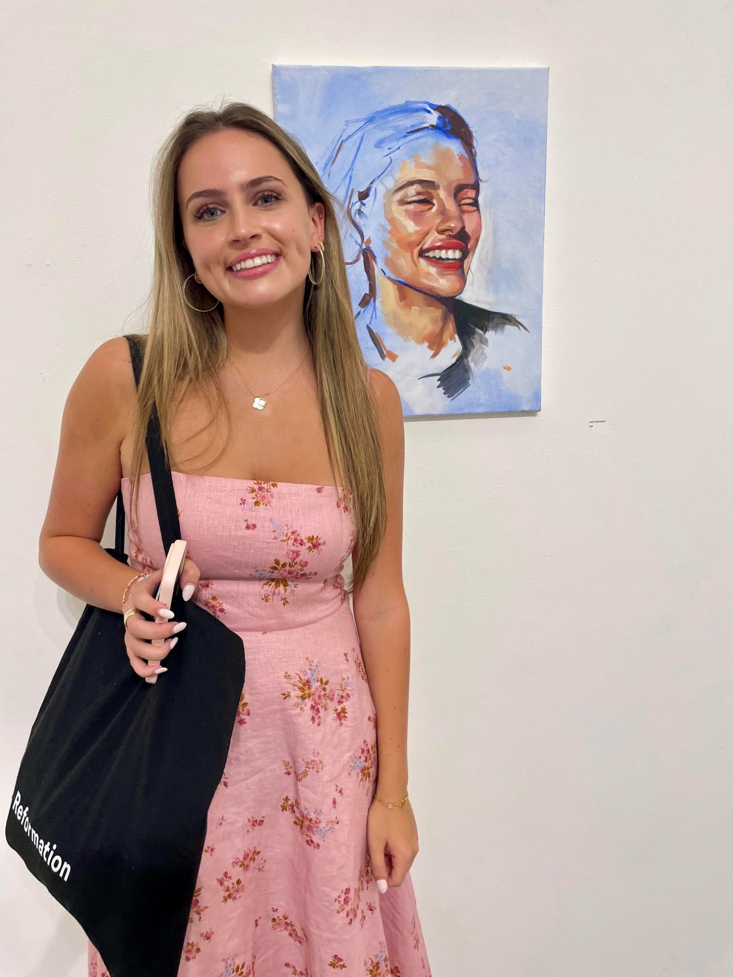 Leila Campbell '25, wearing a pink strapless dress with a black handbag slung over her right shoulder, poses with one of her paintings hung on the wall behind her.