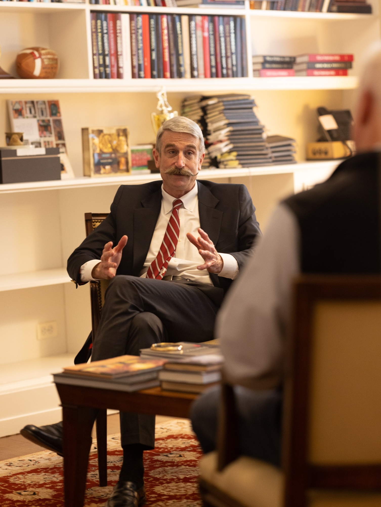 Steve Murray sitting in his office gestures with his hands while talking to another man. He is wearing a blue blazer, white oxford button-down short, and a red and black tie, and dark gray slacks with his legs crossed.