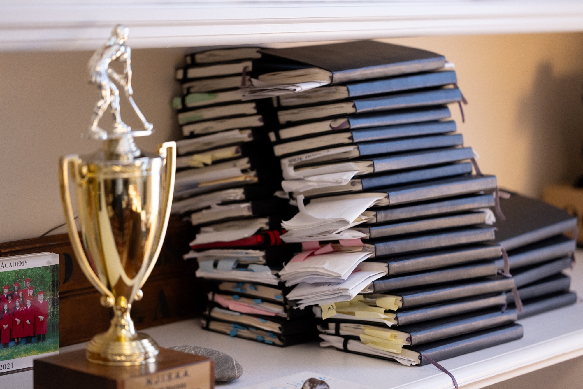 Two stacks of black, hardcover notebooks with papers sticking out lean against a bookshelf wall.