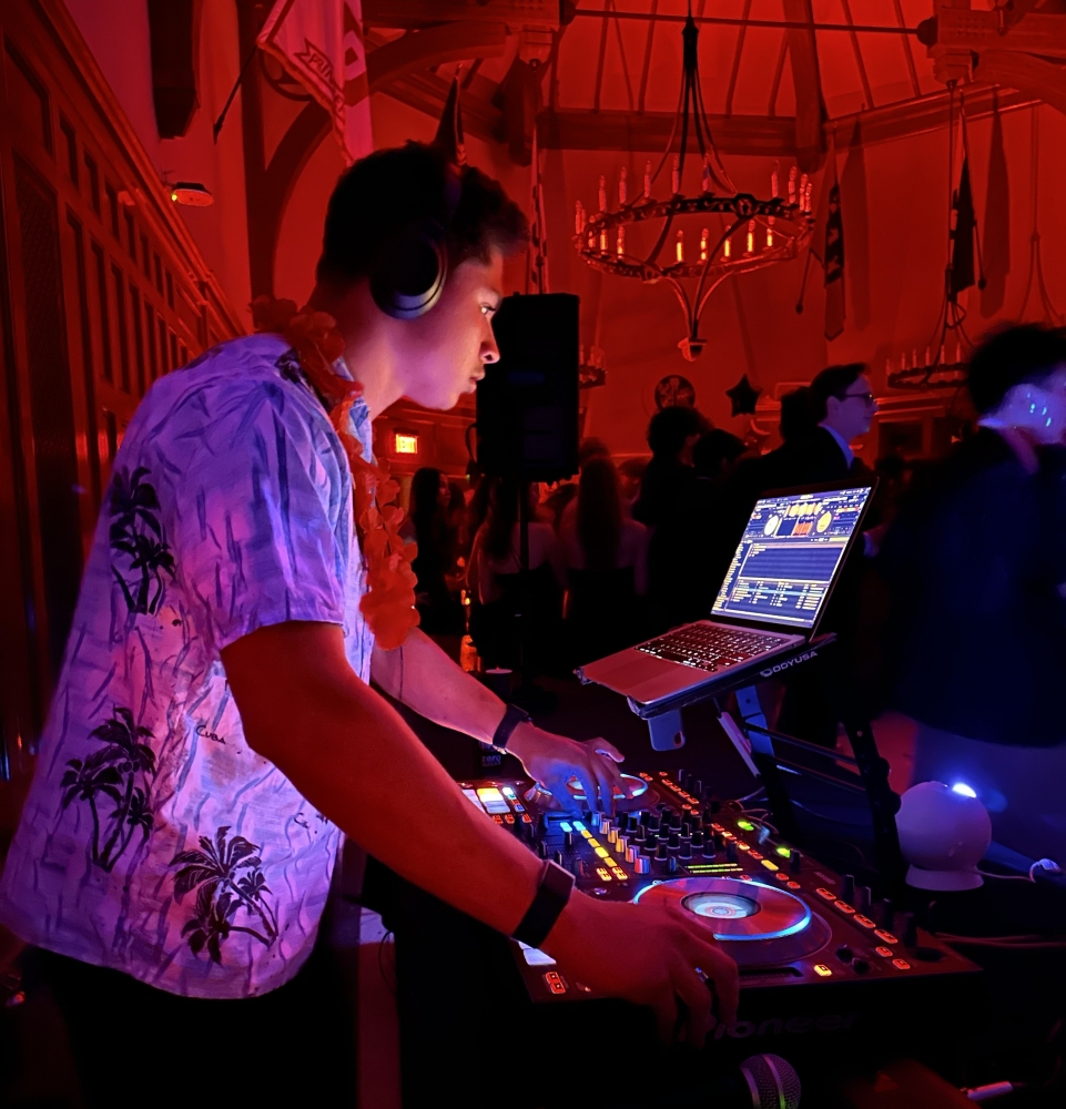 In a room lit red and darkened for a student dance, Alex Salgado-Lozhkin '26 deejays at his equipment board while wearing headphones