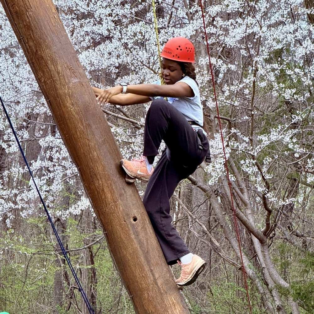 A girl wearing an orange helmet scales a telephone pole-style log leaning upward to the left.