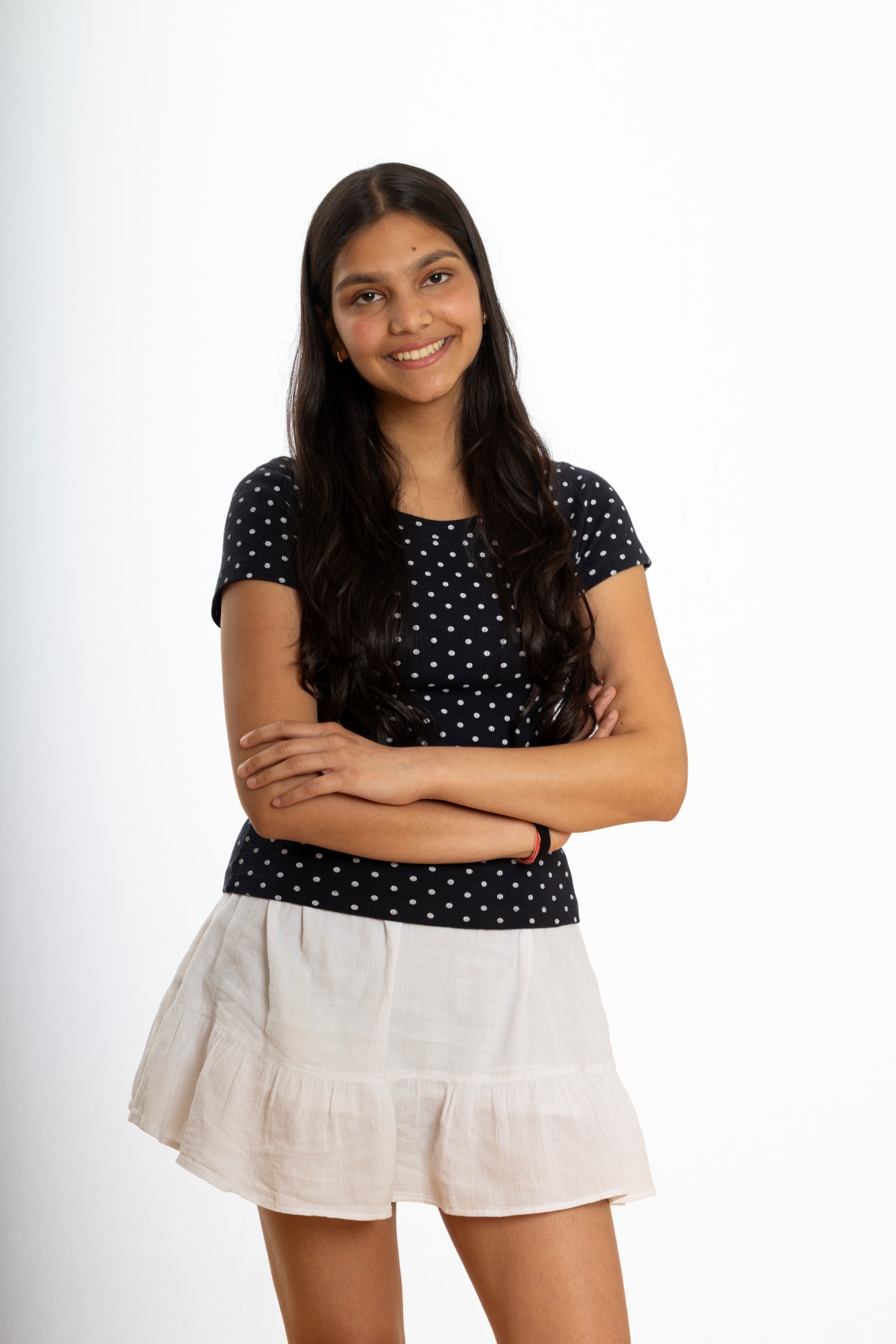 A young girl with long black hair is smiling. She is standing against a plain white background, wearing a black short-sleeve shirt and a white skirt.