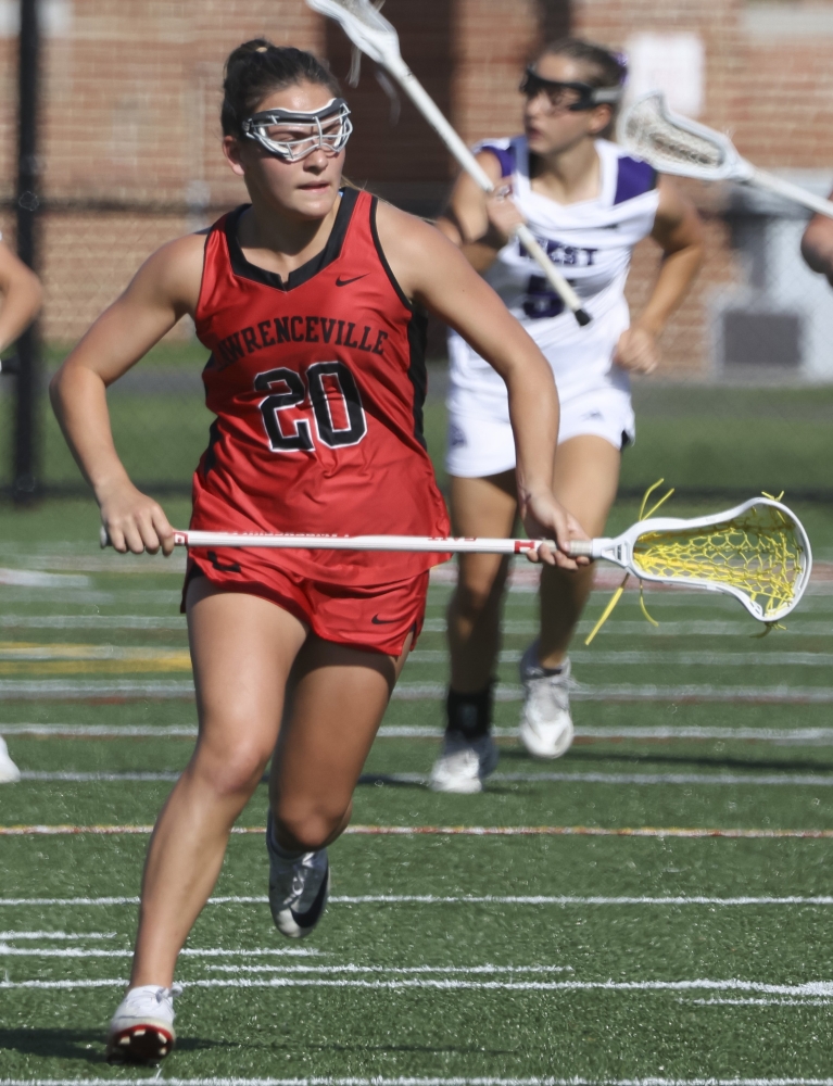 A girl in a red Lawrenceville lacrosse uniform pursues the play while carrying her stick, with the basket to the left.